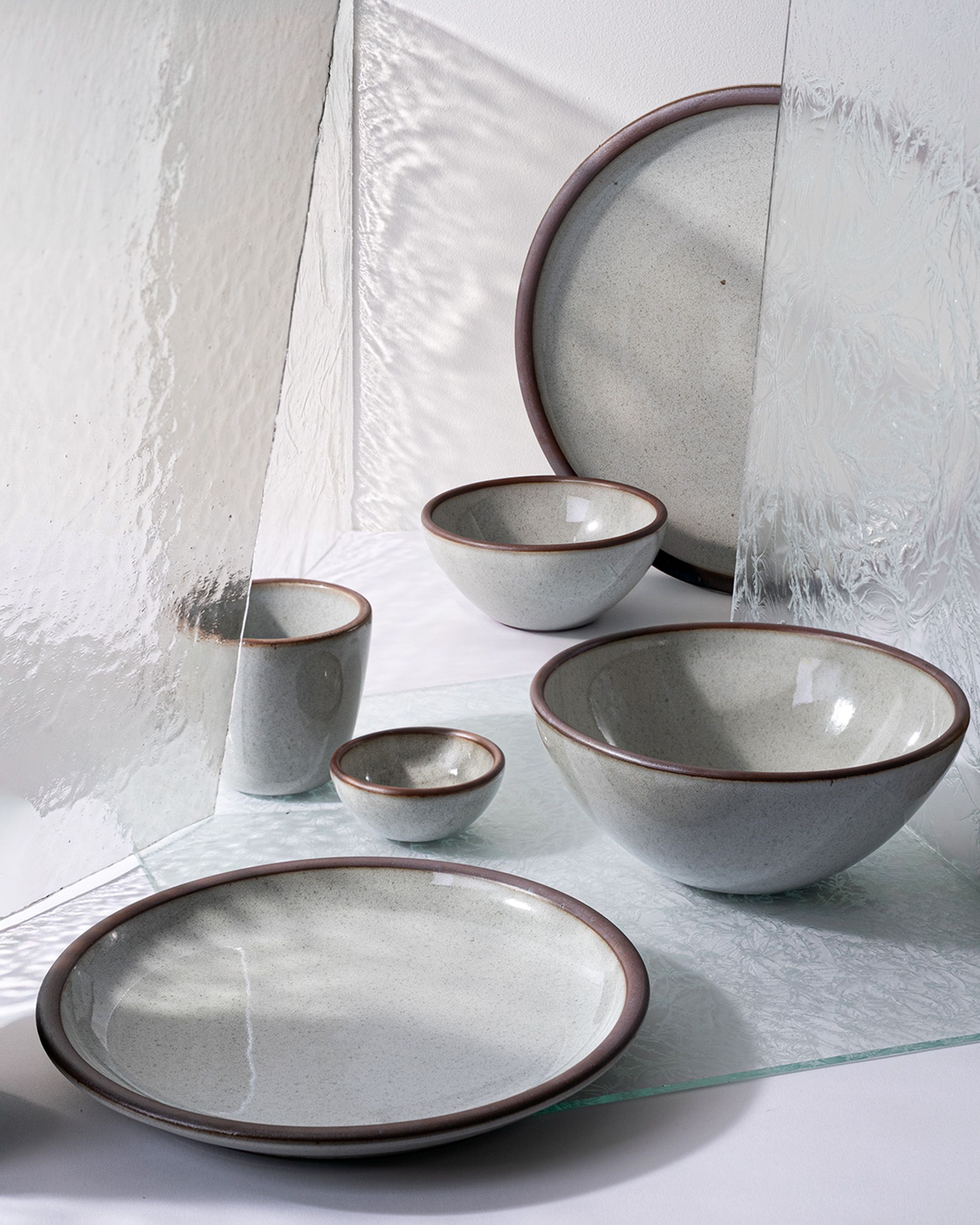 A collection of ceramic dishes in reflective icy white with unglazed rims arranged among textured glass panels, creating reflective light and shadow.