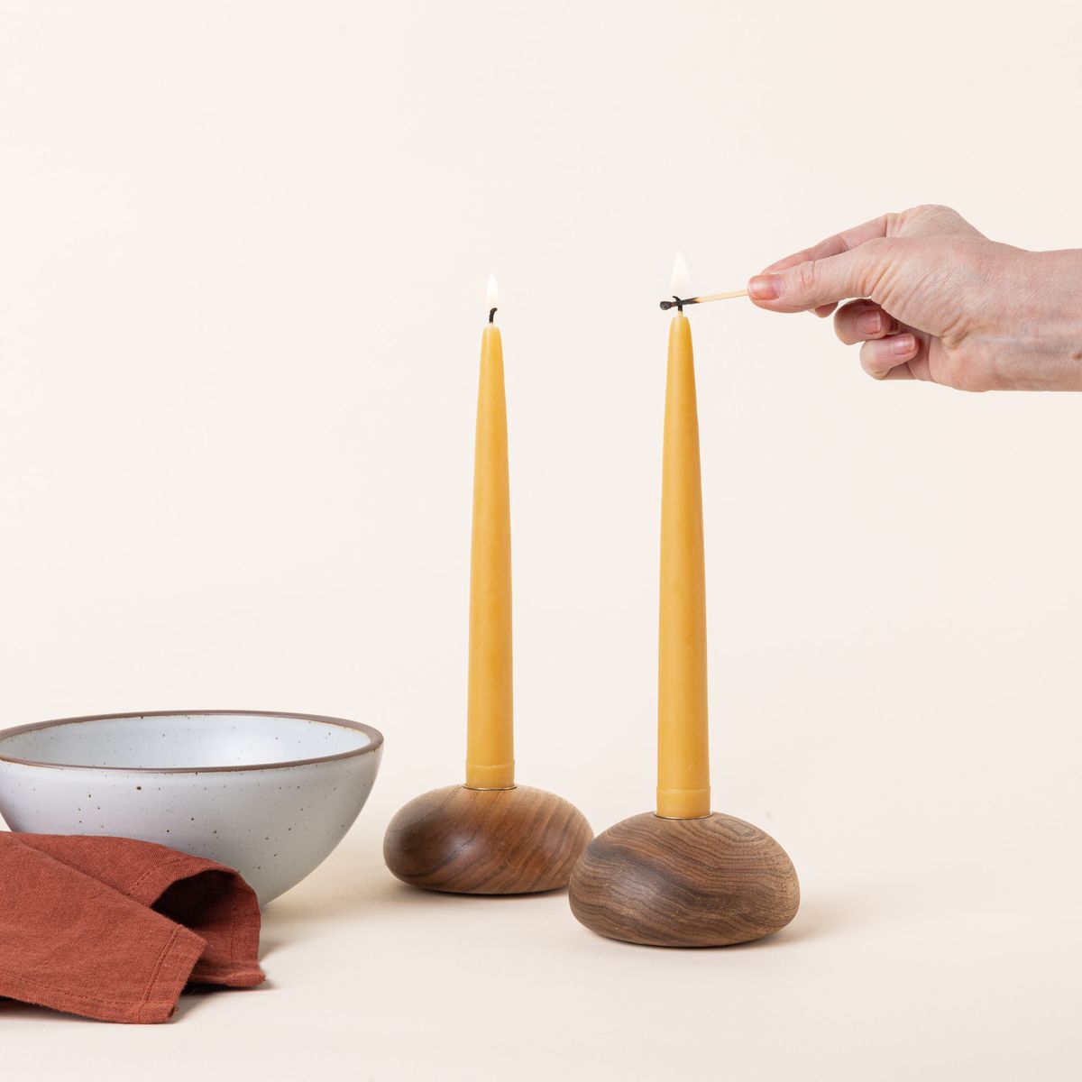 A hand lights a pair of yellow beeswax taper candles sitting in round wooden candle holders