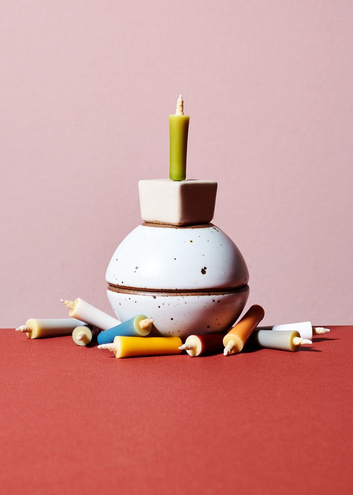 Small, rainbow colored candles on a table. Two bitty bowls stacked ontop of one another, topped with a cubicle tan candle stand with green candle in it.