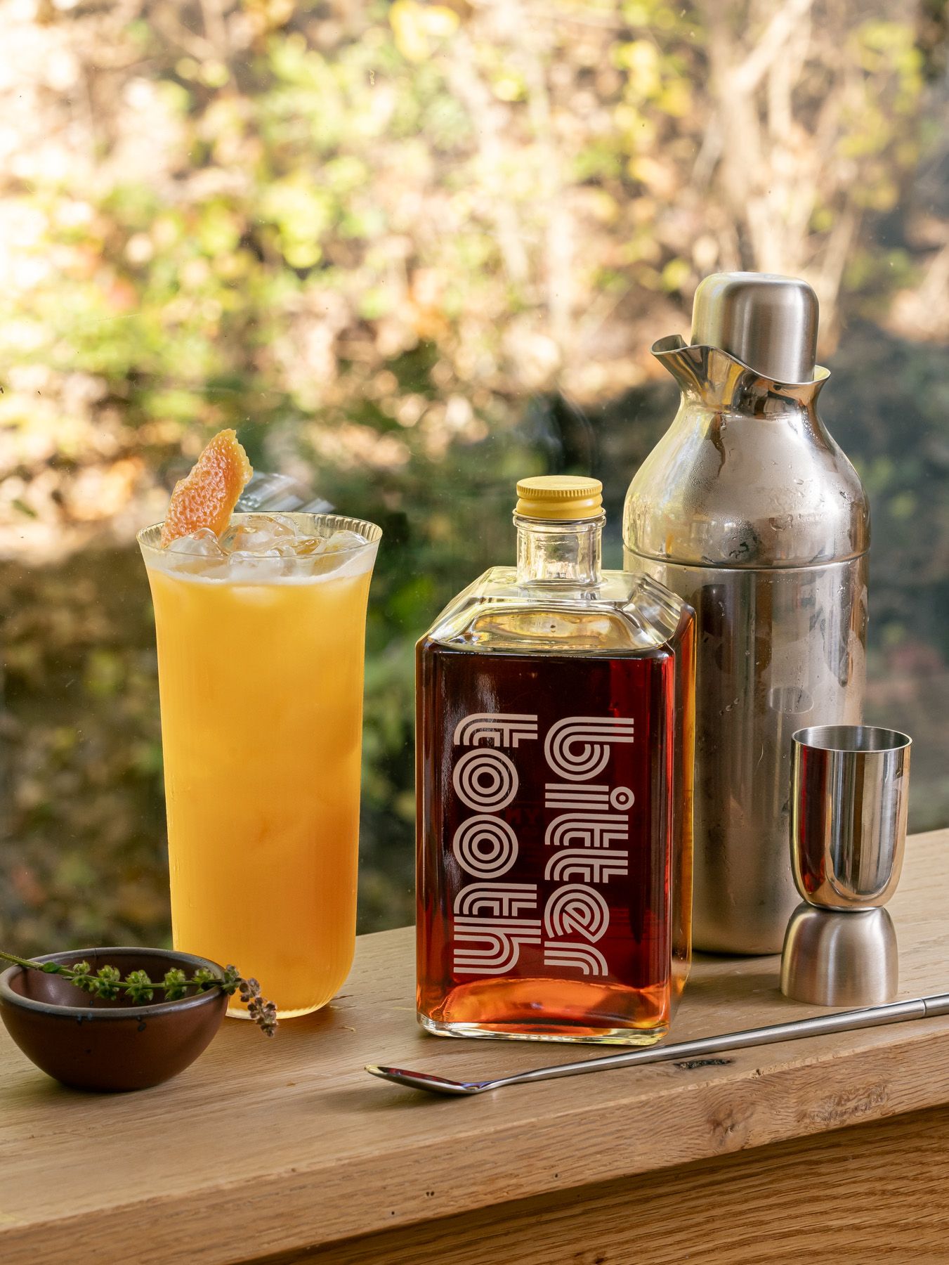 On a window sill sits a tall ribbed glass with a yellow cocktail, a bottle named 'bitter tooth' and a stainless steel cocktail shaker.