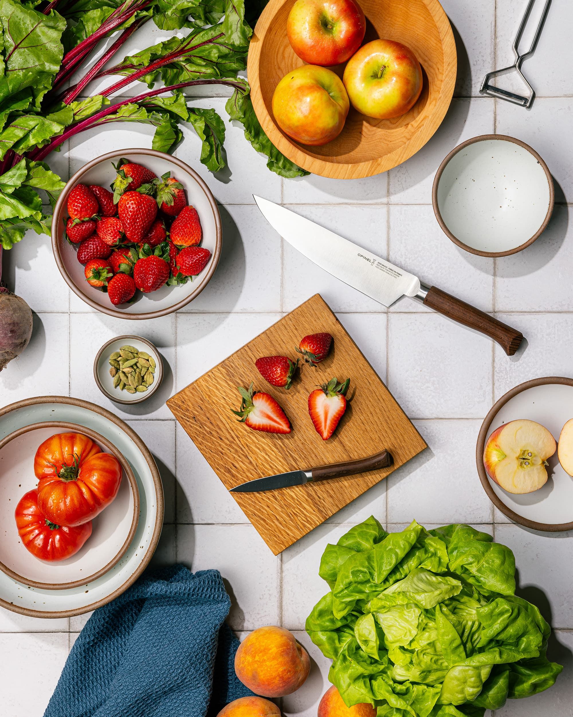 An overhead view of a kitchen counter spread featuring fruits and vegetables being prepped and sliced, chef's knife, paring knife, ceramic bowls and plates in cool white colors, a blue kitchen towel, a small cutting board and more.