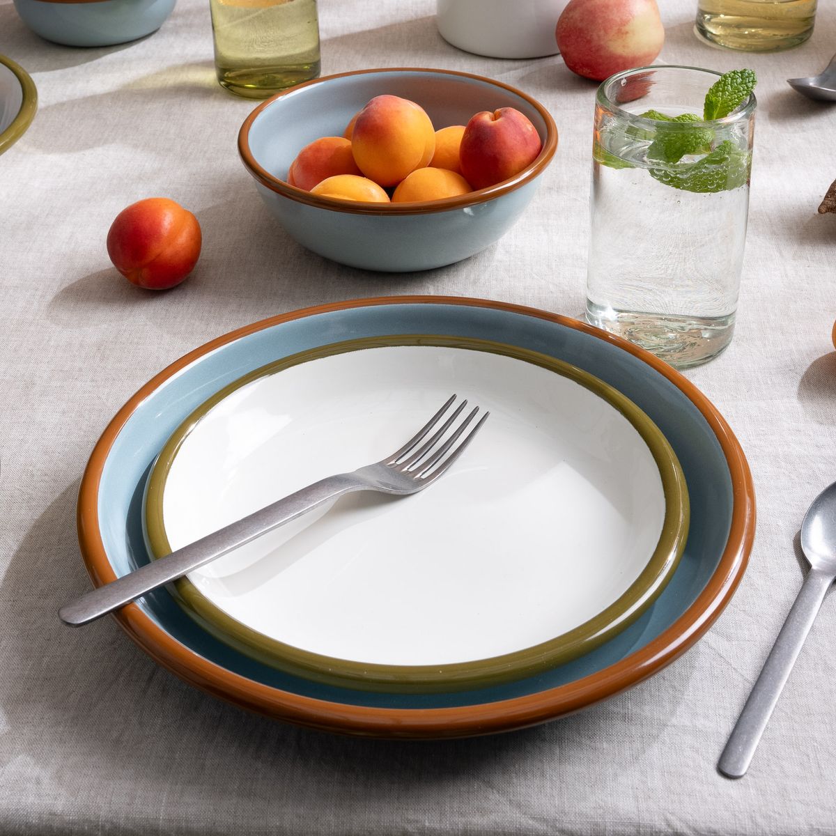 A nestled set of enamel side plate in white and a dinner plate in robin's egg blue on a table with silverware and more.