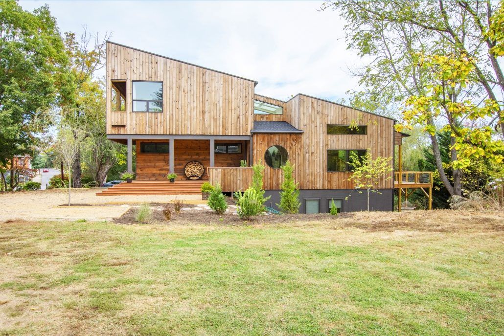 An exterior look at a house with a slanted diagonal roof, wood exterior walls, minimal landscaping, large yard and surrounding trees.