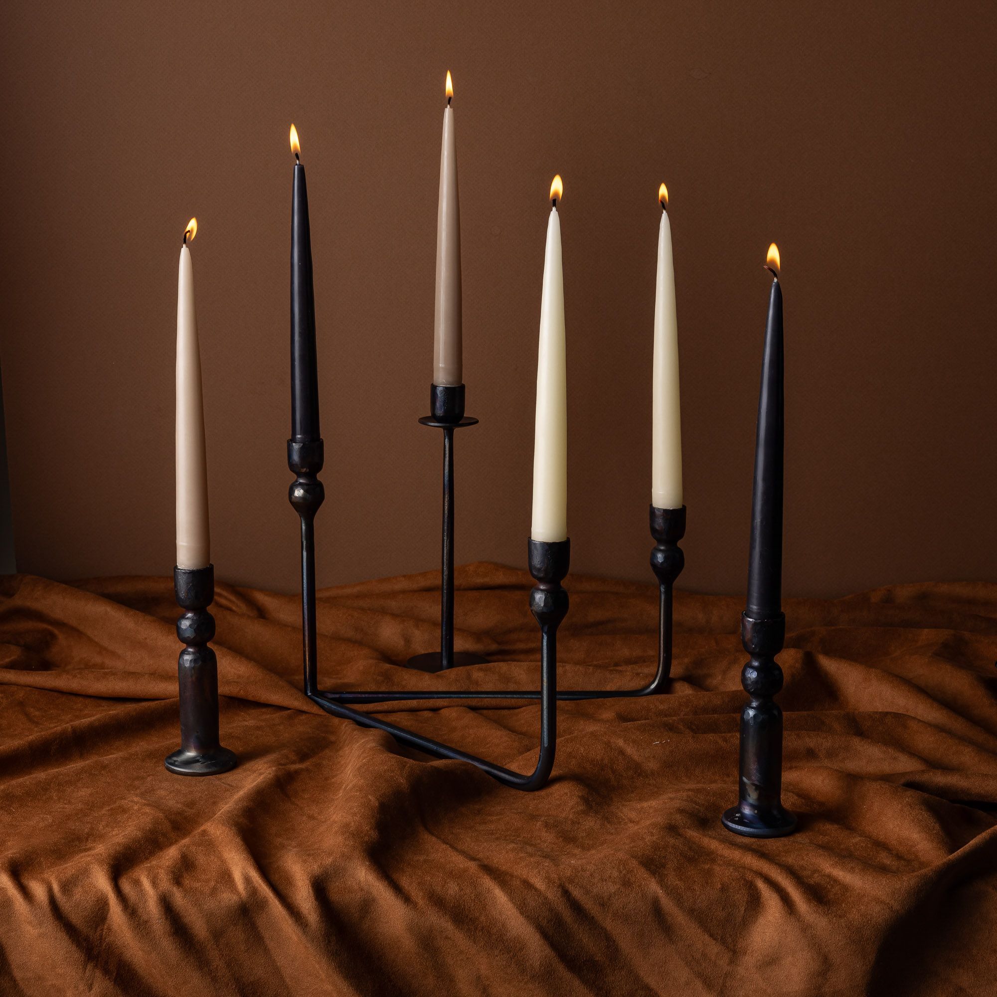 An assortment of iron candlestick holders and a candelabra all holding lit tapered candles. They all sit on a brown fabric with a brown background.