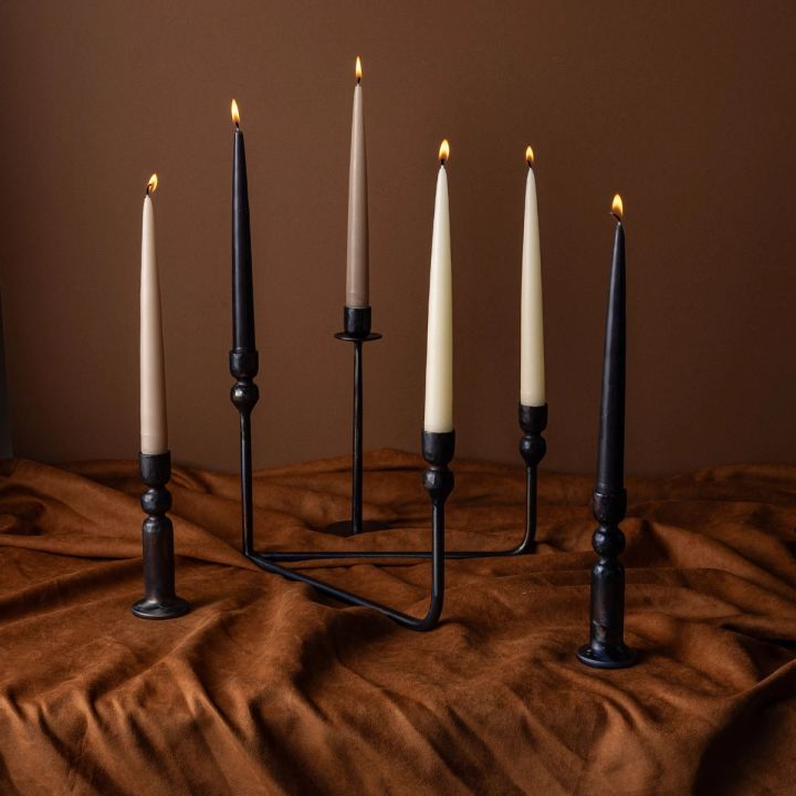 An assortment of iron candlestick holders and a candelabra all holding lit tapered candles. They all sit on a brown fabric with a brown background.