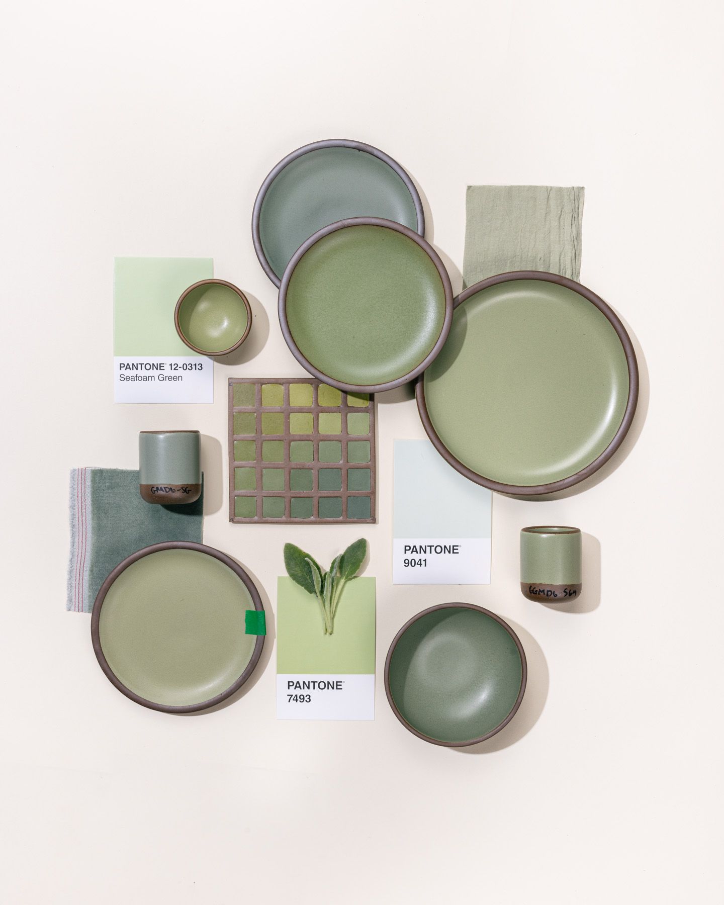 An overhead view of a flat lay featuring 2 ceramic plates in varying calming sage colors, surrounded by pantone color cards, illustrations, and a ceramic color tile also all in sage green tones.