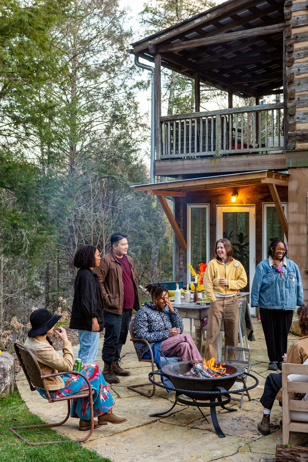 A gathering of people around a campfire outside a two story cabin.