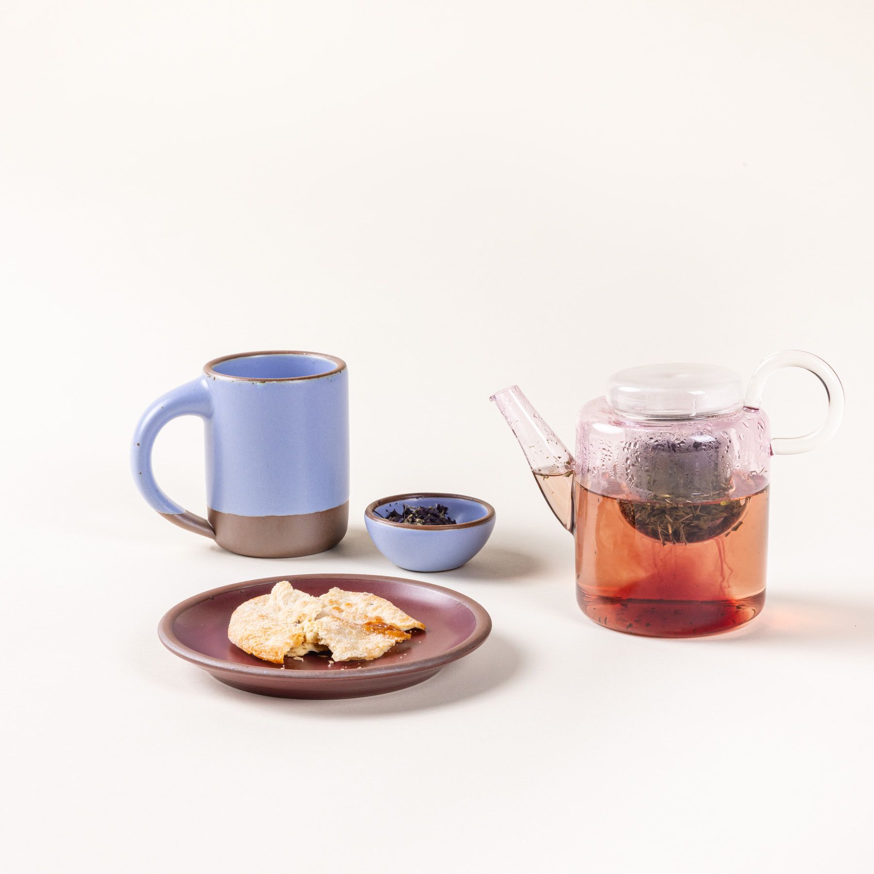 A modern glass teapot in a faint pink color with tea inside sits with a ceramic mug and small breakfast