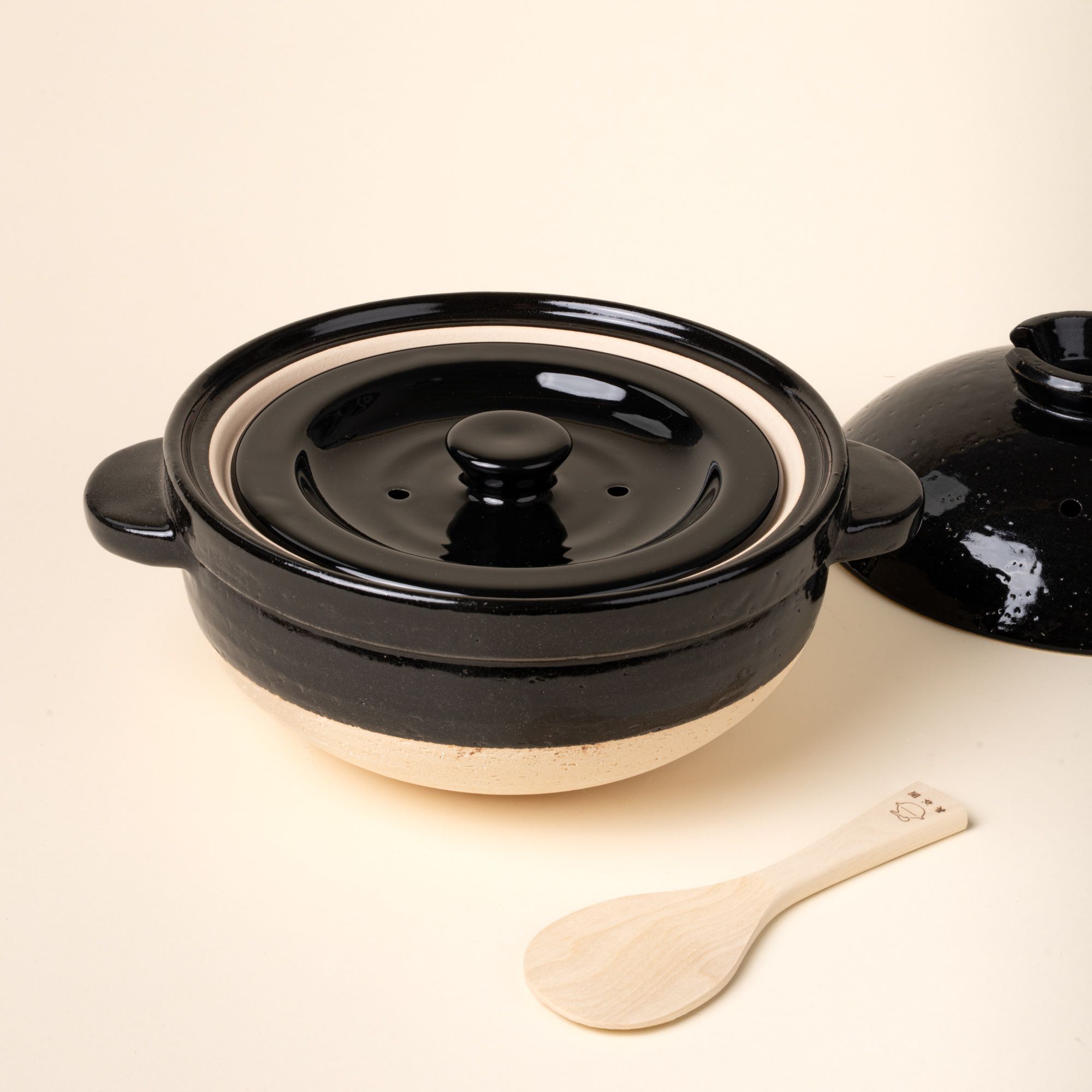 Black ceramic donabe pot with lid and a small wooden spoon on a light background.