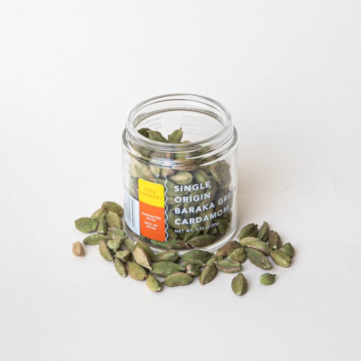 Open glass jar filled and surrounded with whole cardamom and a transparent label that reads "Single Origin Baraka Green Cardamom"