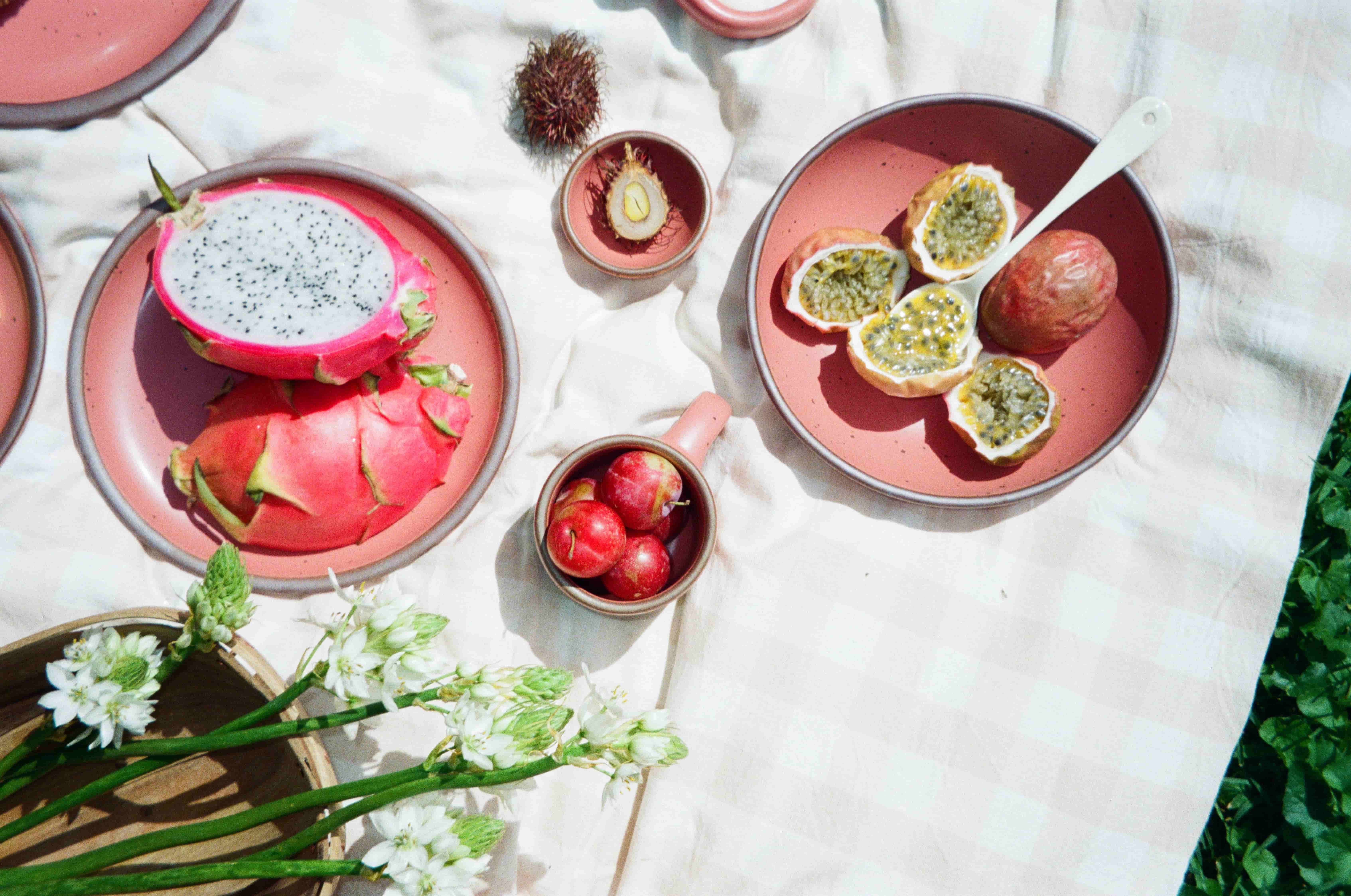 Halved dragon fruit, passionfruit, and plums on watermelon pink plates atop a cream picnic blanket, with white flowers and striped napkin.