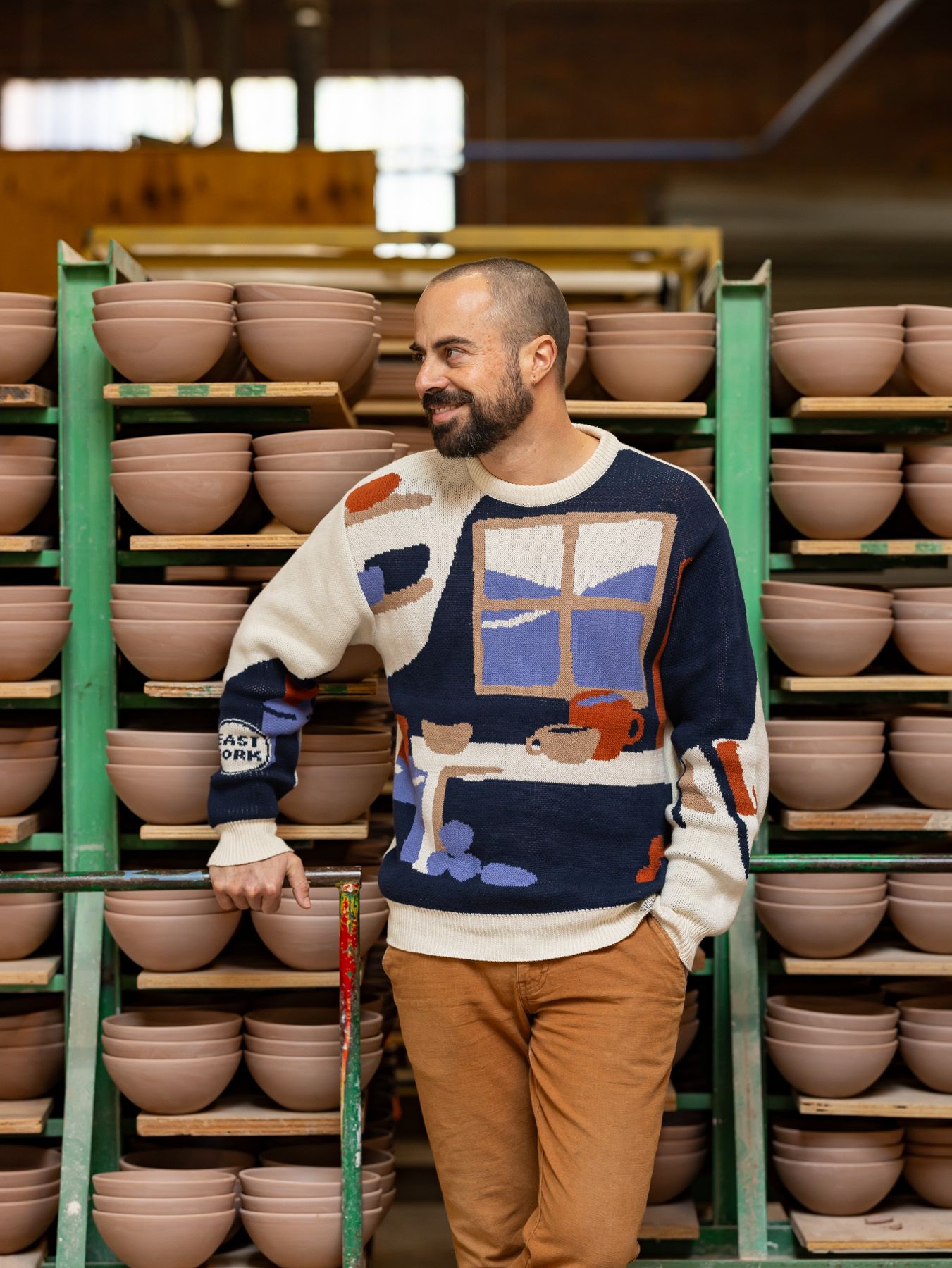 Alex wears a sweater with an illustration of a pottery workshop with a large window, shelves, and table with mugs and bowls. Alex stands in front of a rack of ceramic bowls in progress.