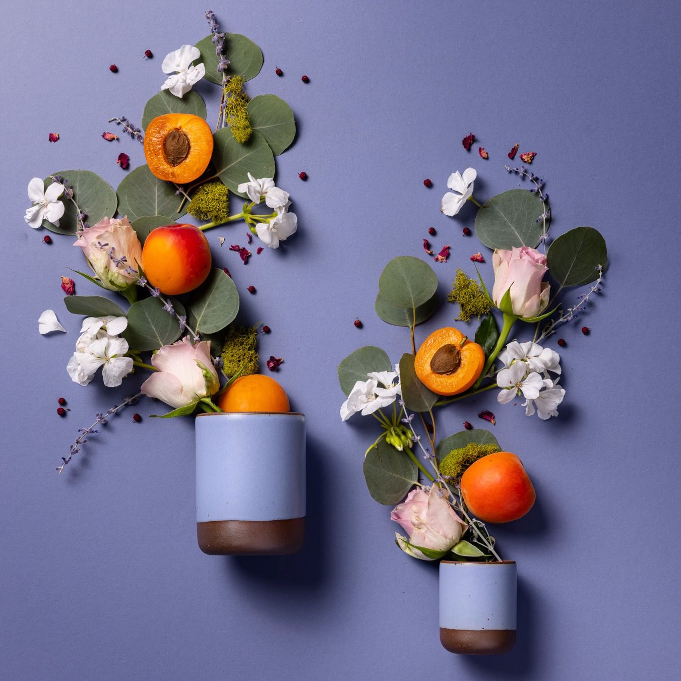 A small and large cylindrical ceramic vessels in a periwinkle color laying on its side - roses, fruits, and greenery are artfully styled coming out of the top of the candles to reflect the scent.