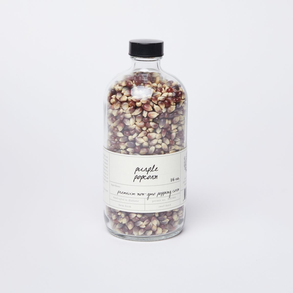 A clear glass bottle full of purple and white popcorn kernels with a black lid and a white label that reads "Purple Popcorn"
