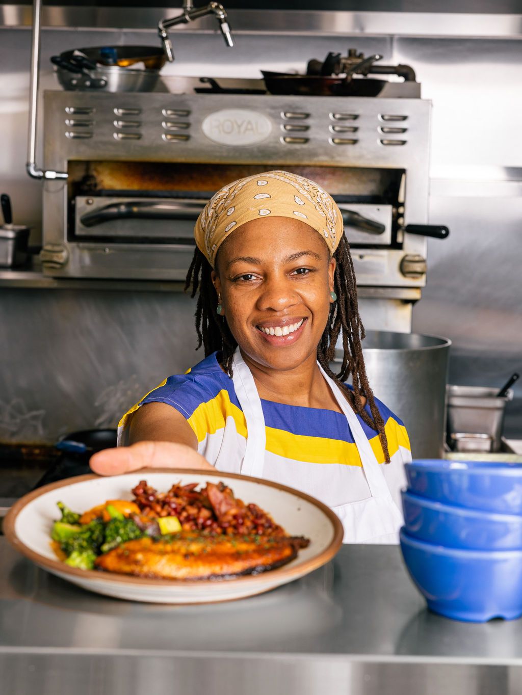 A woman presents a plate of food from a restaurant kitchen and is smiling.