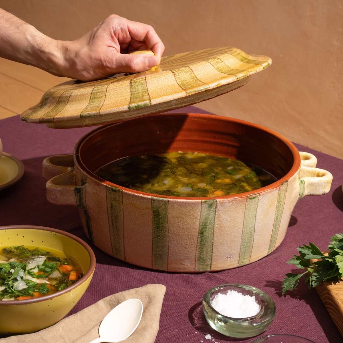 A hand holds a lid above a striped green and yellow casserole dish filled with soup.