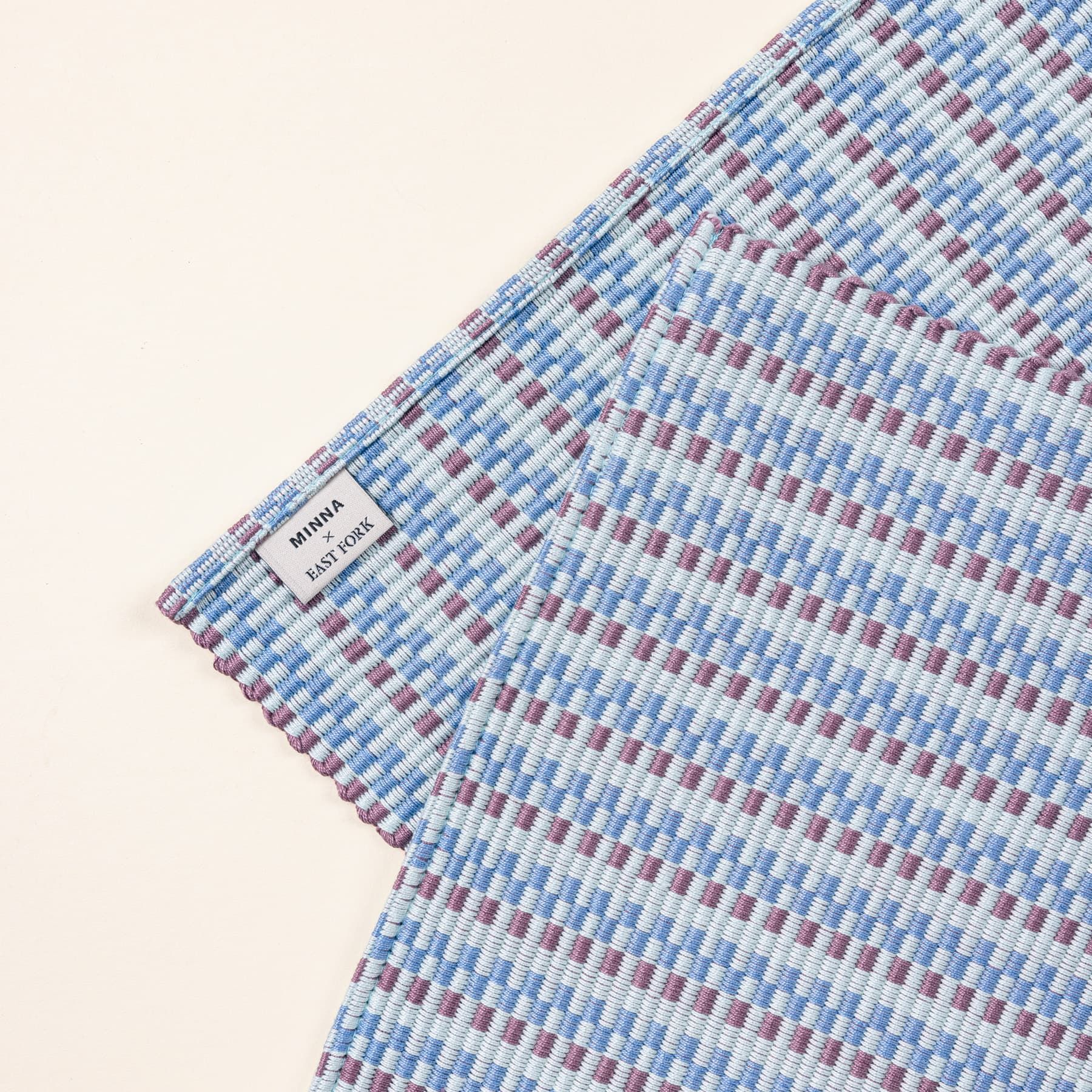 A close up of a woven table runner with thin horizontal striped soft blues and purple colors. A tag is on the back corner and reads 'Minna x East Fork'