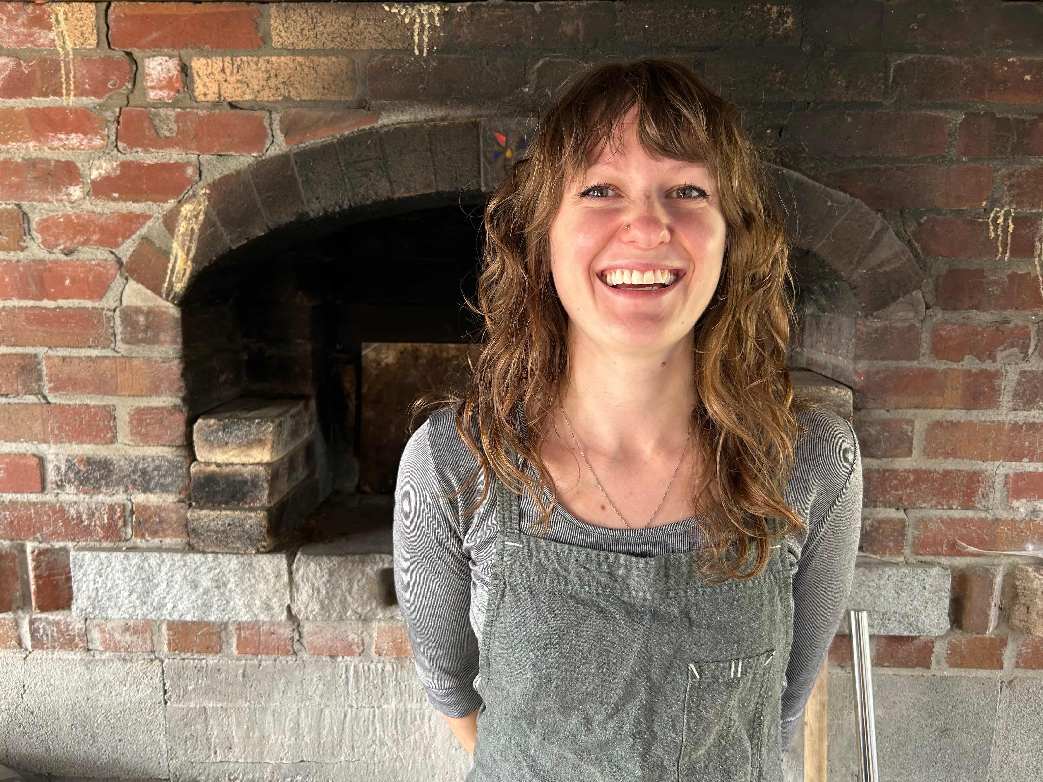 A photo of Camille smiling and wearing an apron in front of a stone fire