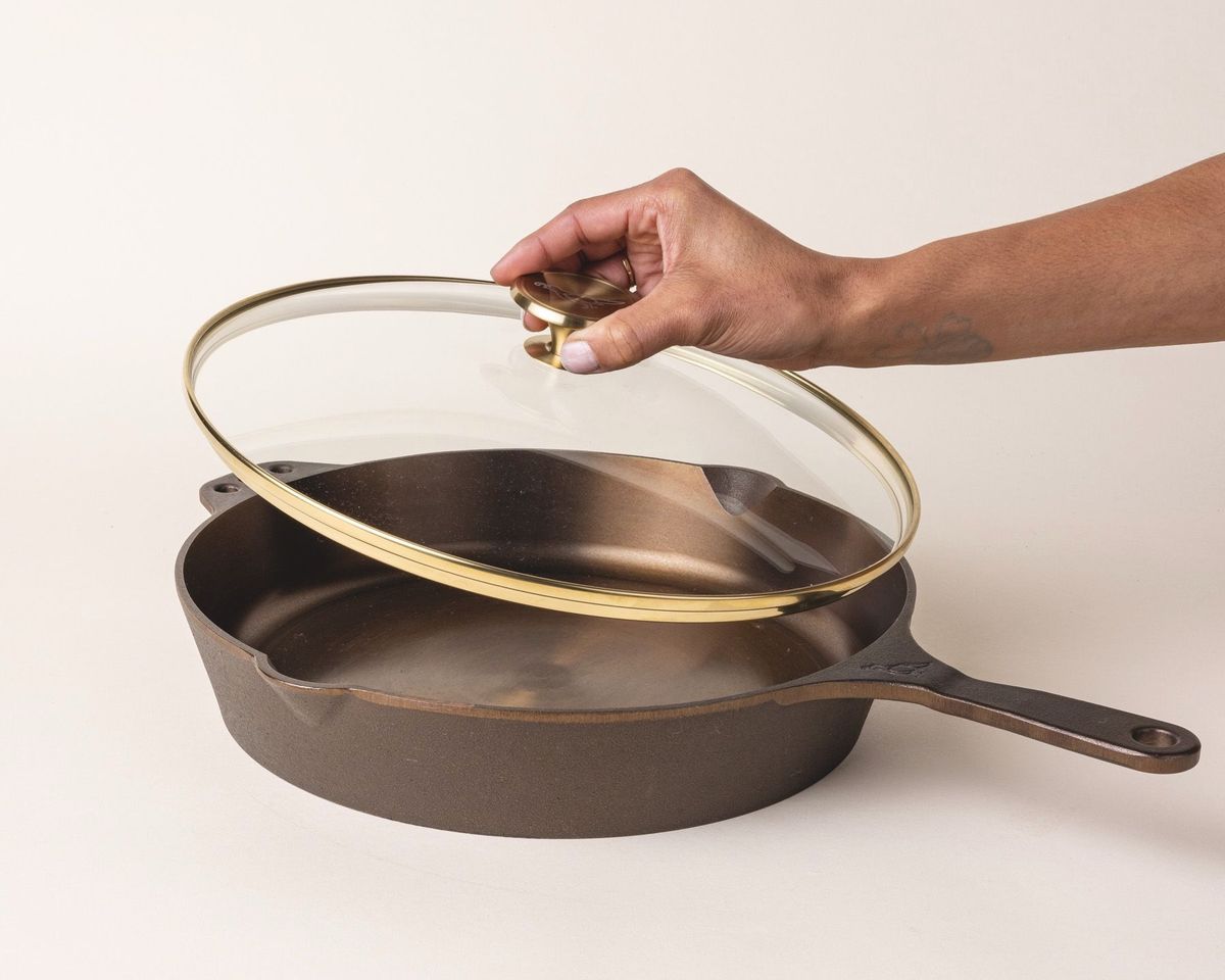 A hand holds up a glass skillet lid with a brass lip and brass handle knob over a cast iron skillet