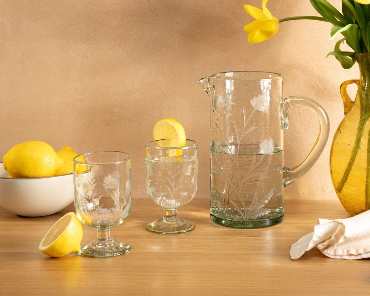 Etched thistle wine glasses and pitcher sit on a wooden table next to a bowl of lemons and vase of tulips.