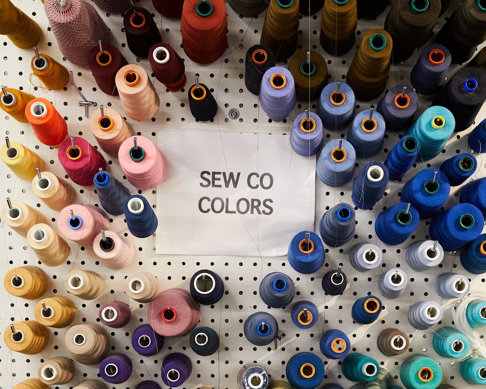 A peg wall filled with large spools of colorful thread and a sign that reads "SEW CO COLORS"