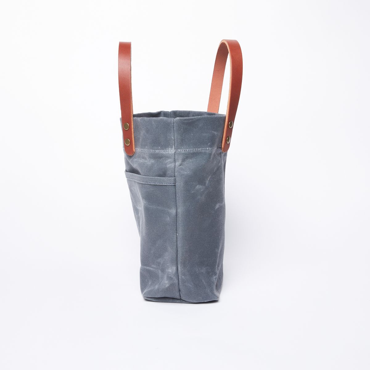 Side view of a blue grey waxed canvas bag with two external pockets on one side and a set of leather handles