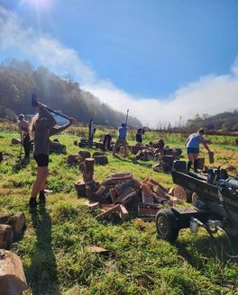 Several people outside chopping wood with the mountains behind them.