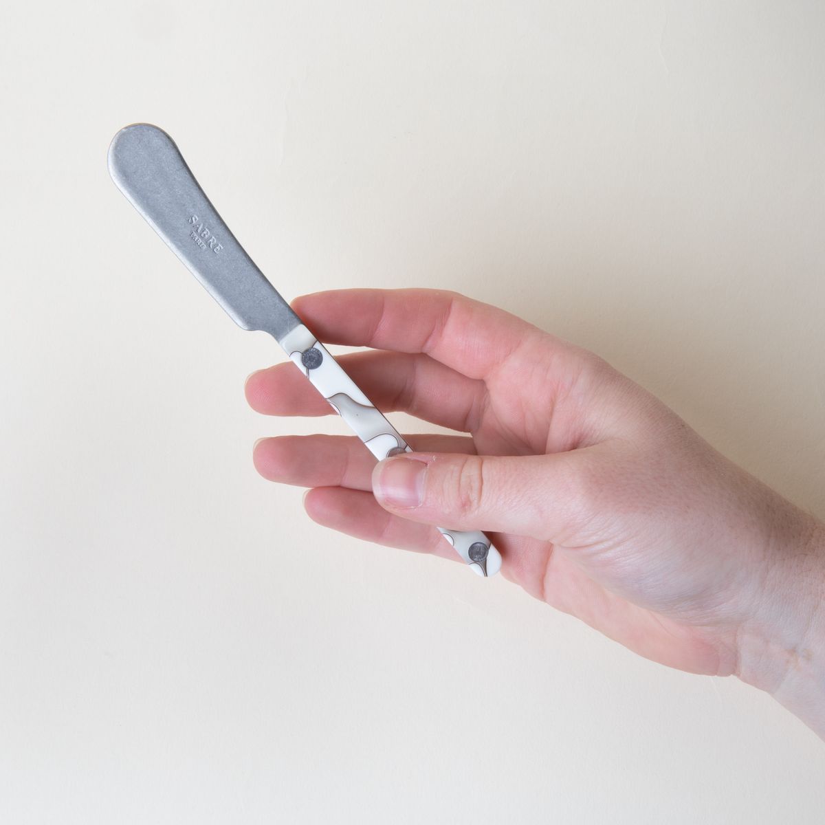 A hand holding a butter knife with a stainless steel blade and an acrylic white and grey handle