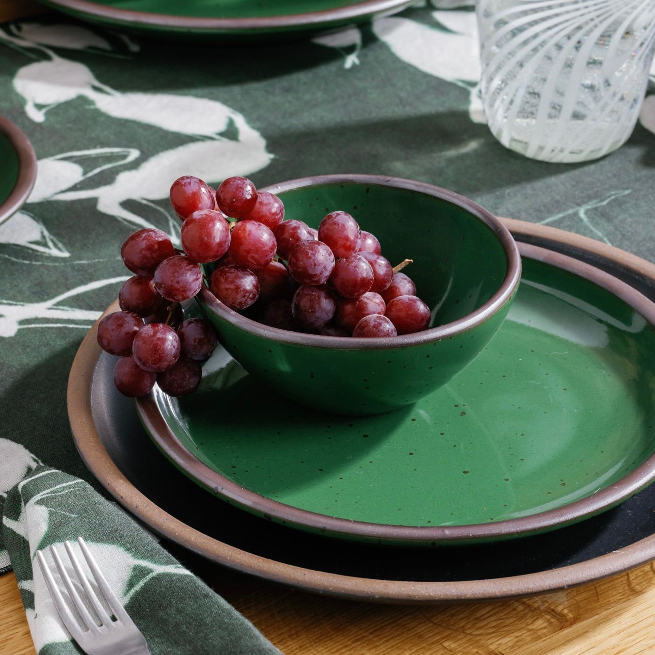 Glossy emerald green ceramic bowl stacked on a matching plate, filled with red grapes, set on a patterned tablecloth with silverware.