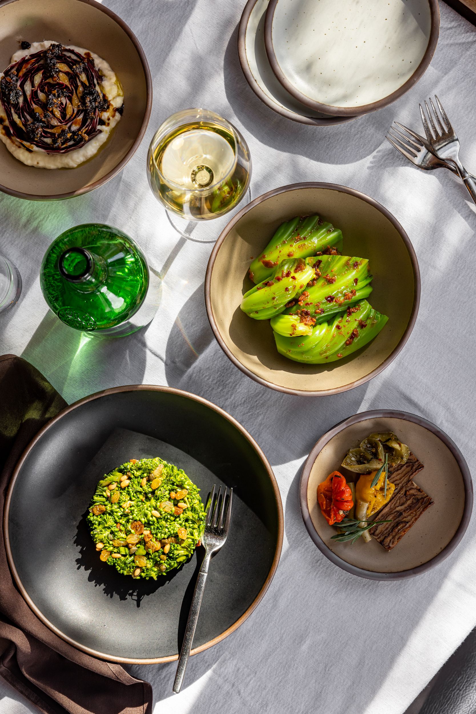 On a table, there are ceramic bowls in black and neutral colors filled with fresh foods, surrounded by a wine glass, green bottle, and silver flatware