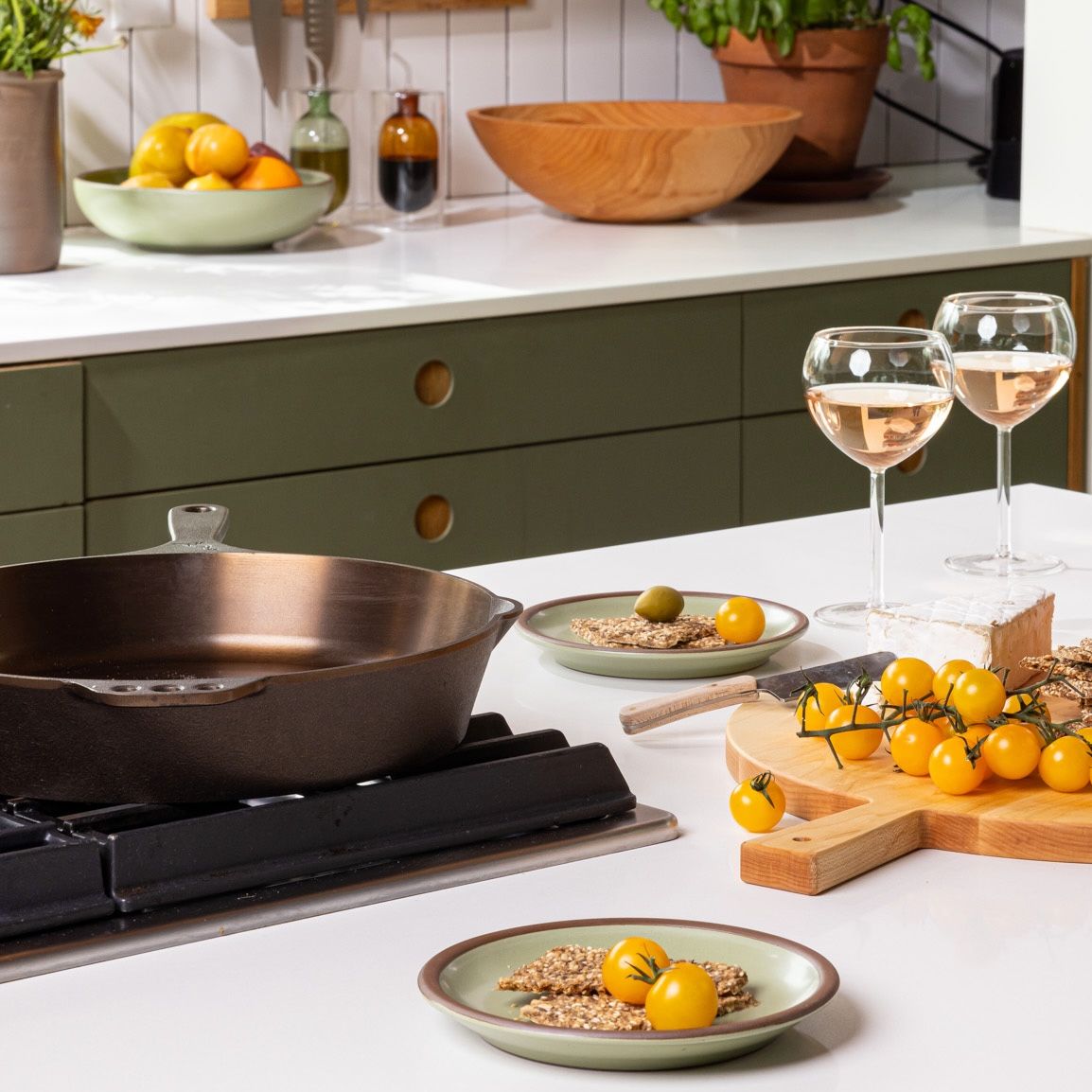 On a kitchen island, sits a cast iron skillet on a stove top, with a charcuterie board with sage green plates and wine glasses nearby.