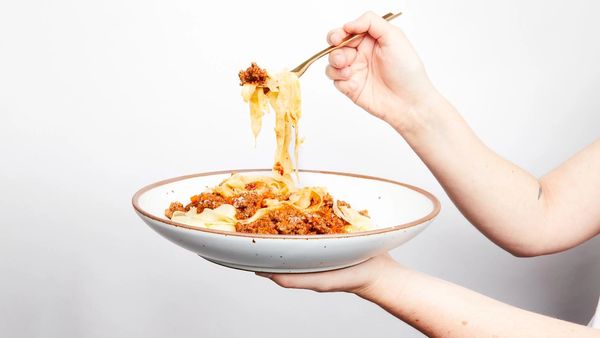A hand holds The Coupe in white, filled with pasta. Another hand holds a fork and is scooping up pasta.