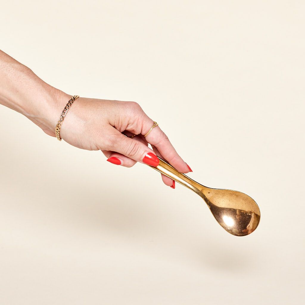 A hand holds a shiny brass spoon, with the back of the spoon's bowl visible