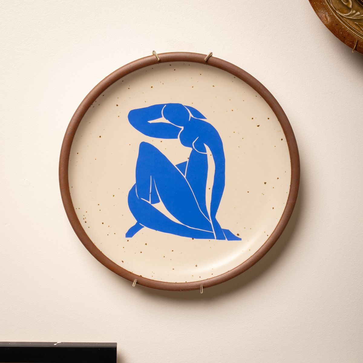 A plate sitting in a brass plate hanger on the wall. The plate has a decal of artwork from Henri's Matisse's Nu Bleu series.