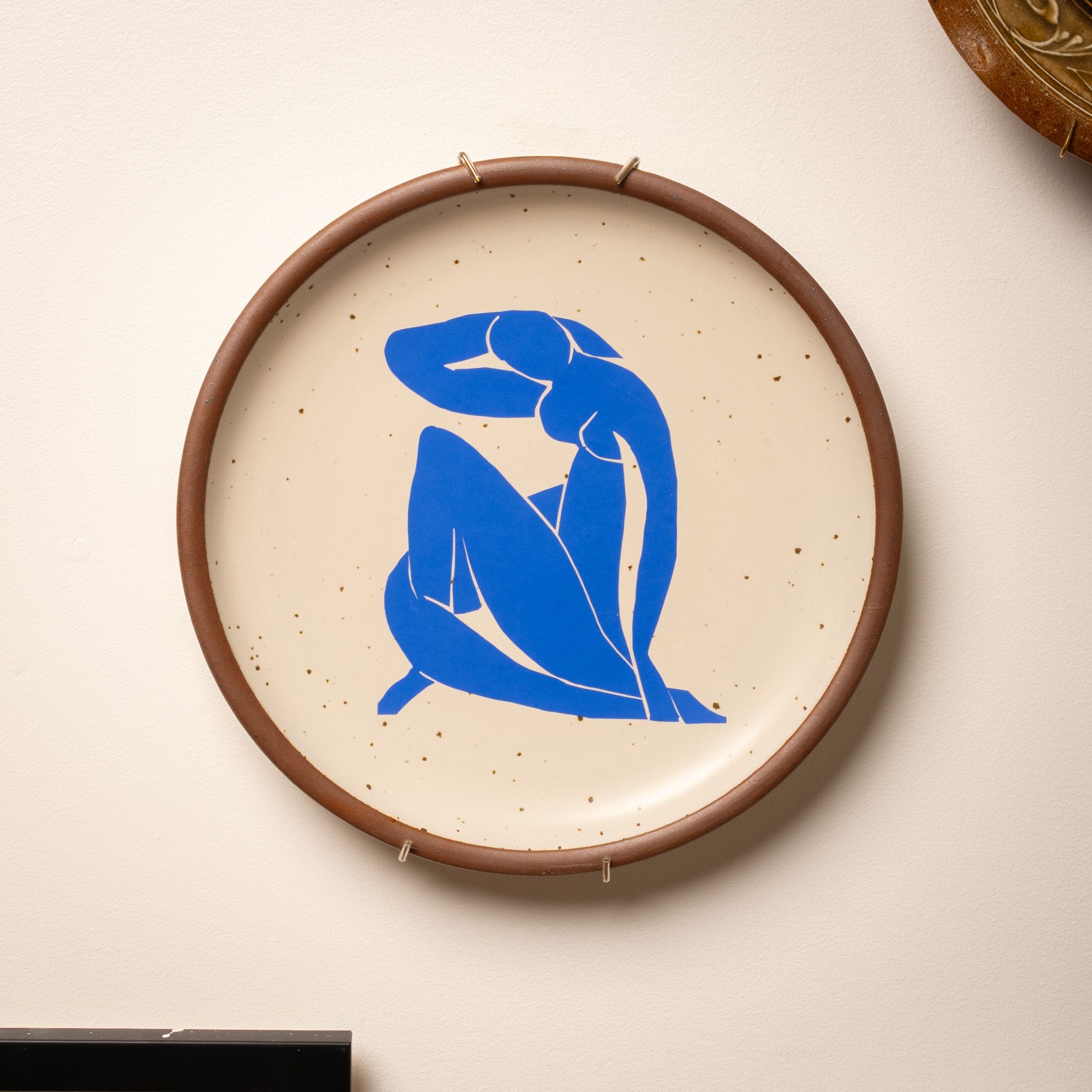A plate sitting in a brass plate hanger on the wall. The plate has a decal of artwork from Henri's Matisse's Nu Bleu series.