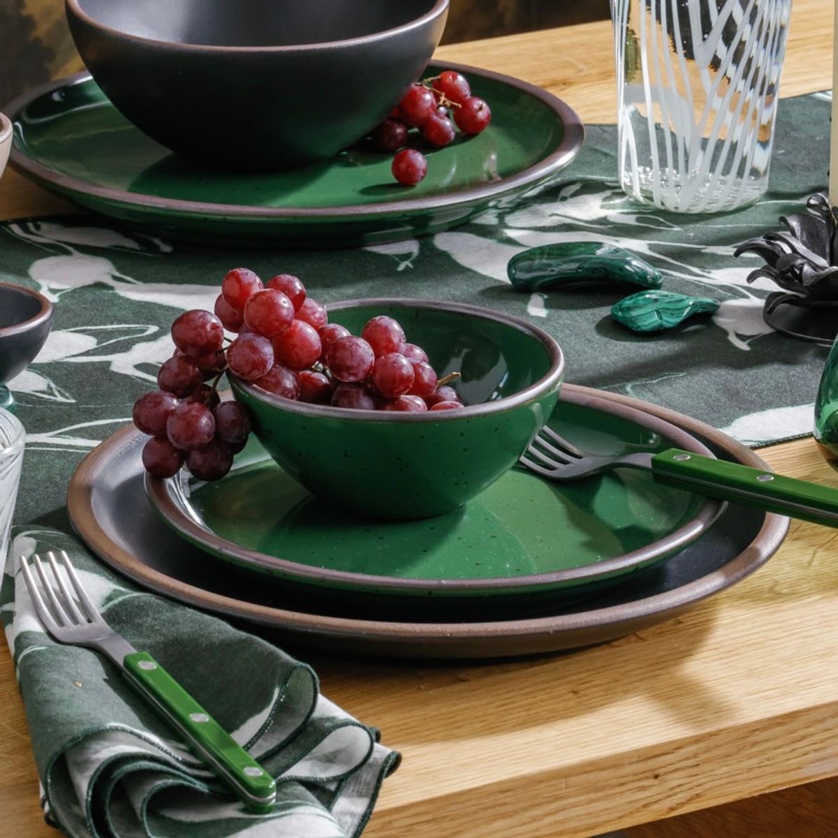 Glossy emerald green ceramic bowl and plate set with grapes on a dining table, paired with a matching green-handled fork and patterned napkin.