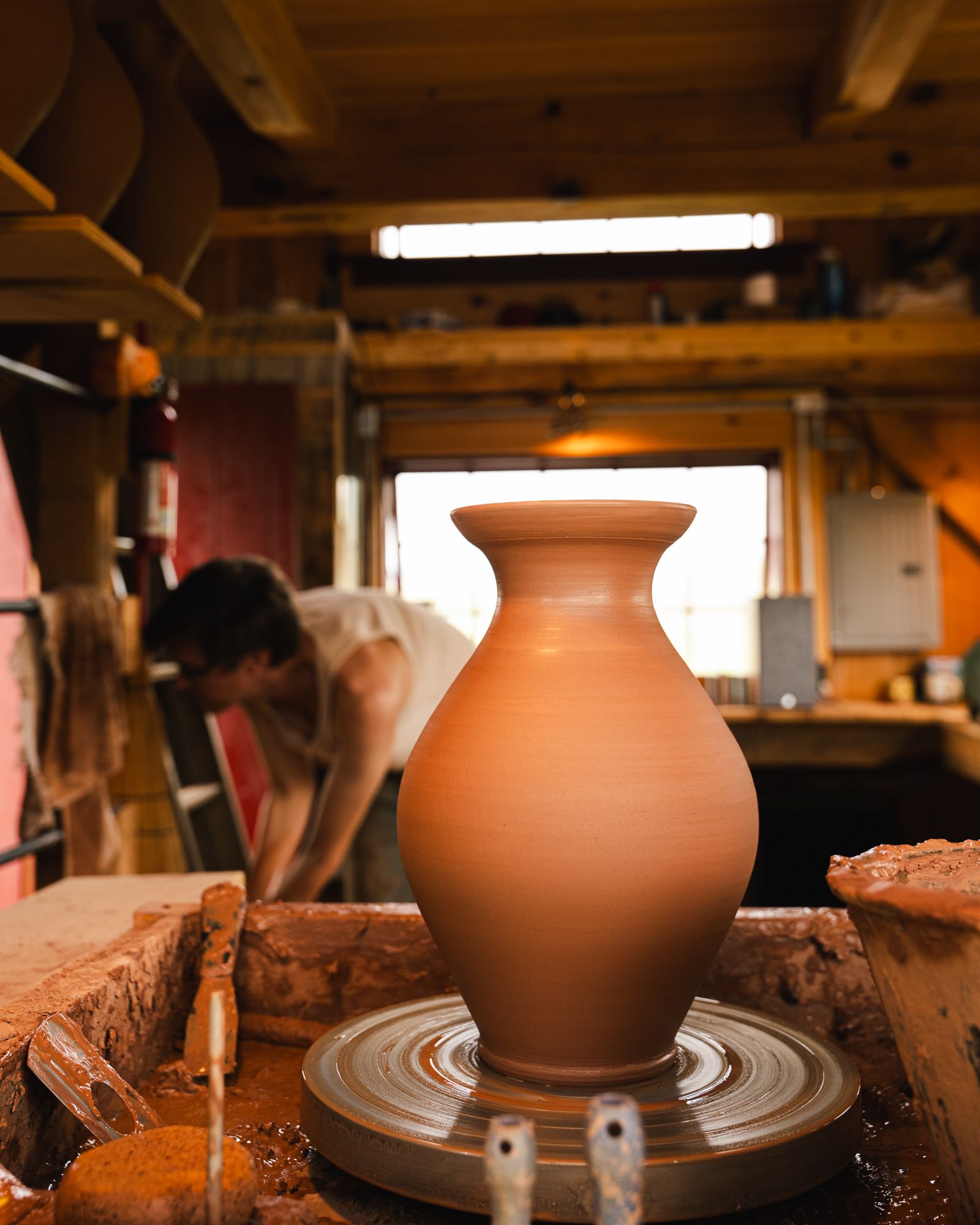 A vase sitting on a potter's wheel with John in the background in his small pottery studio.