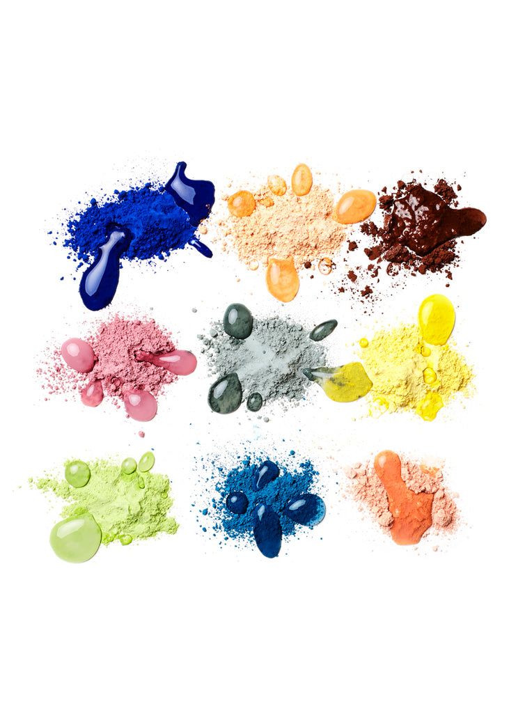 Powdered pigments in individual piles of indigo, orange, pink, gray, yellow, green, blue and peach, each with drops of water that create circles or ovals of the same color