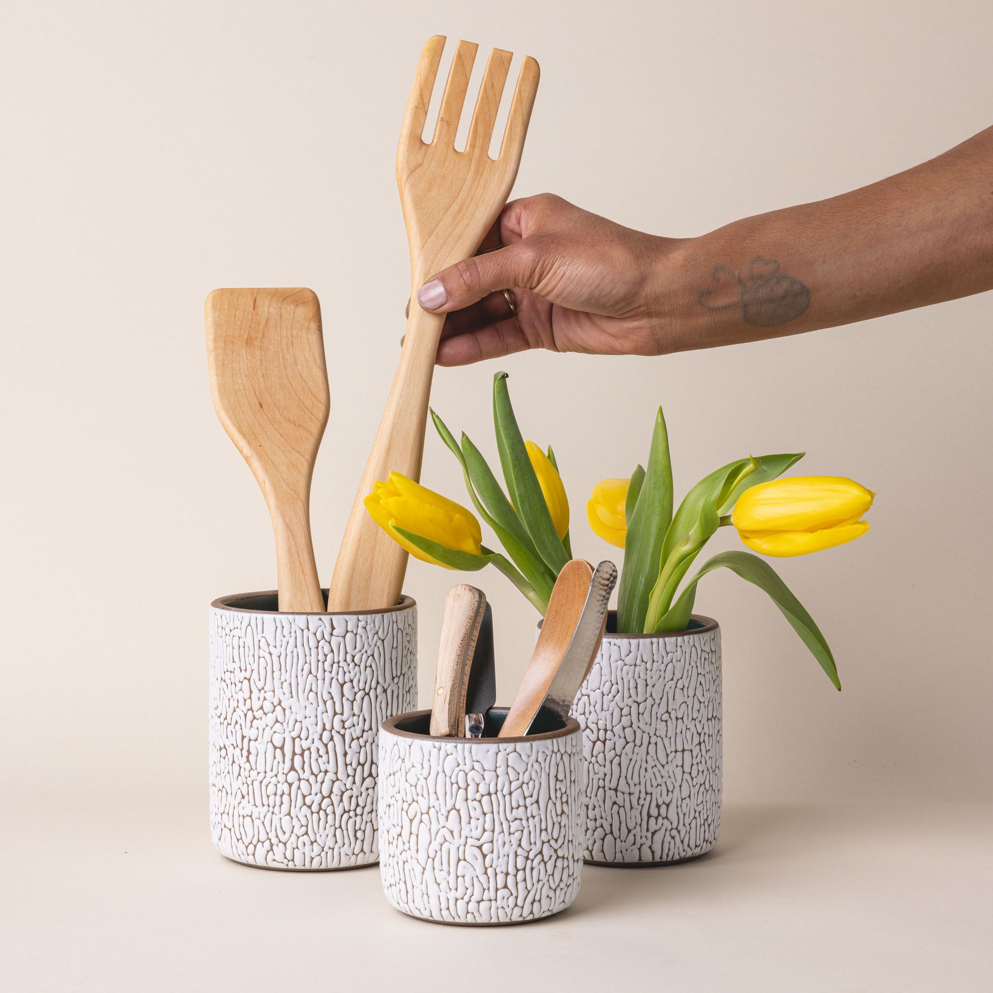 Three white ceramic vessels in Small, Medium, and Big with cracked texture and the interior being dark teal. Each vessel is filled with wooden utensils, flowers, flatware.
