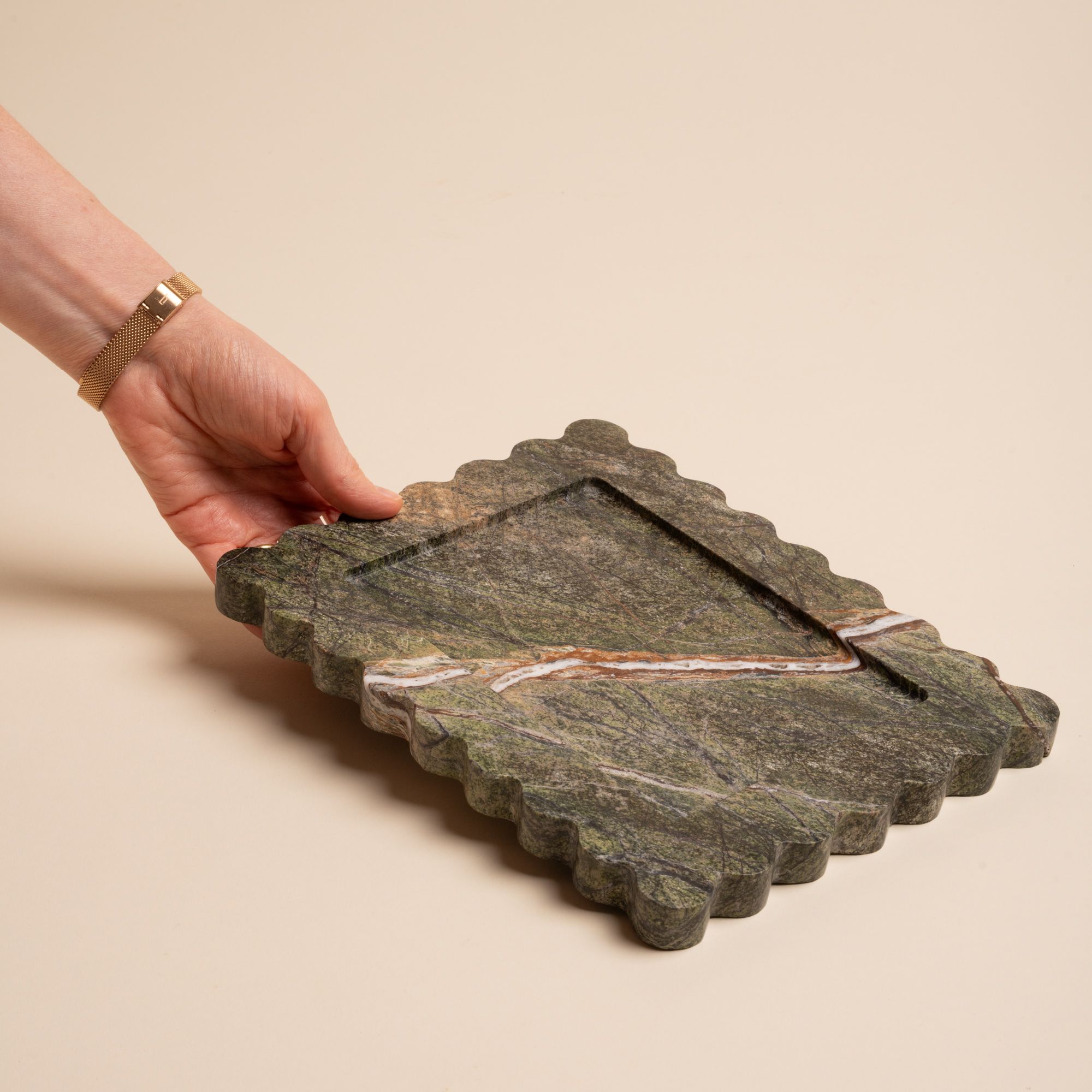 A hand holds up a rectangular green marble tray with scalloped edges and a bold diagonal vein of white and rust running across its surface.