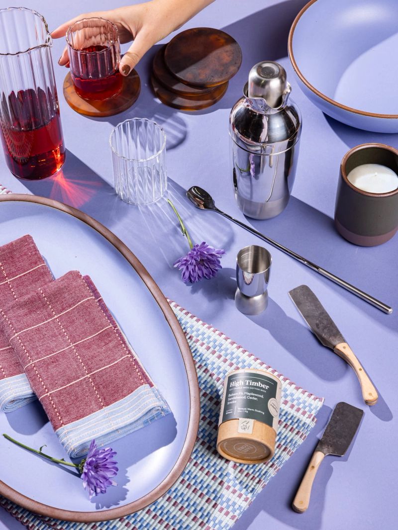 A collection of gifts including a cocktail shaker set, glassware, a mug, a large oval platter, and more against a periwinkle background.