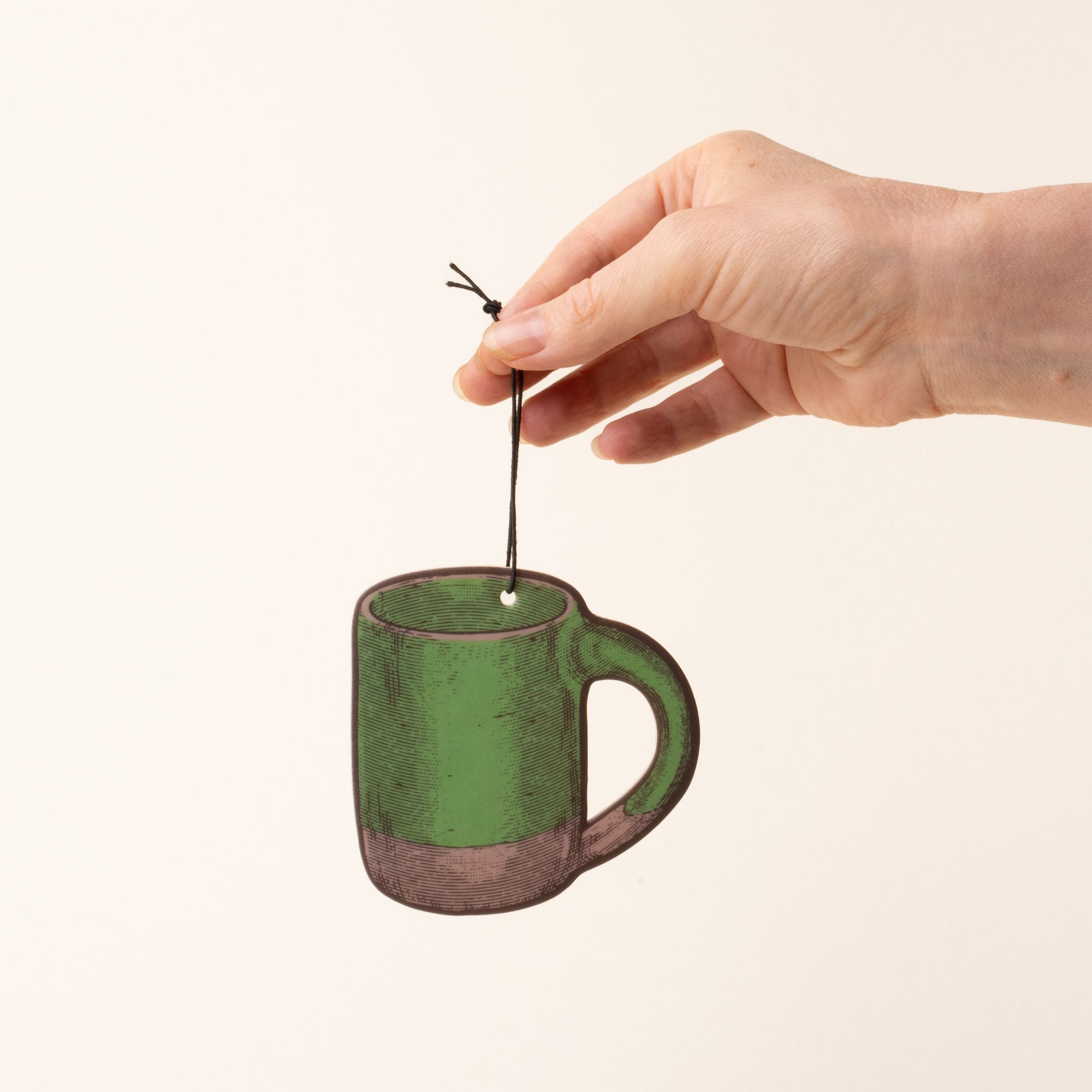 A hand holds an illustrated mug-shaped air freshener in a green color with a black hanging string against a light cream background.
