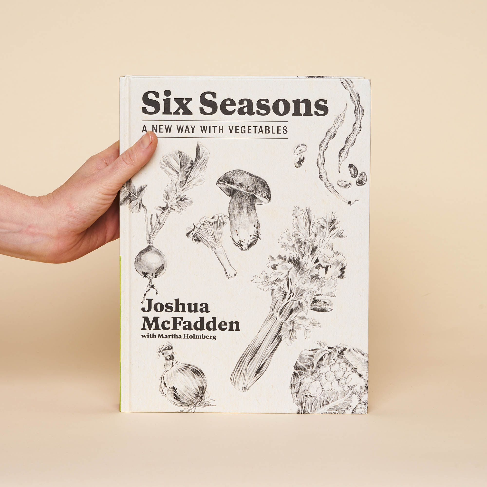 A hand holds an off-white cookbook with delicate illustrations of beets, onions, mushrooms, green beans and celery