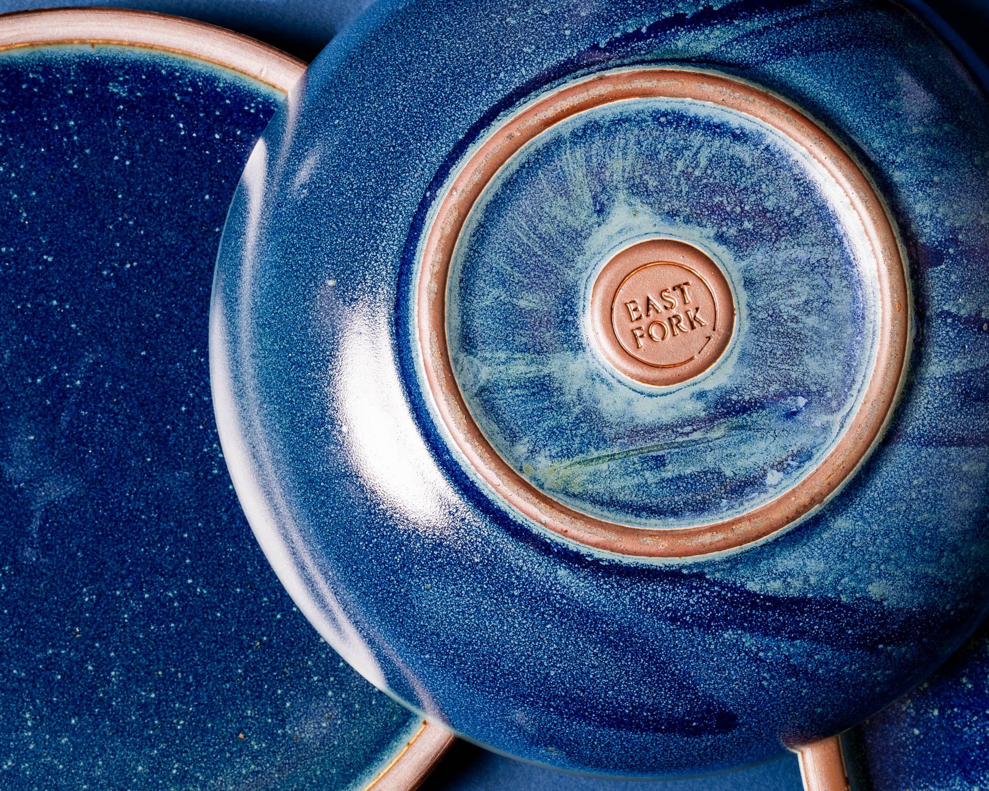 A shallow ceramic bowl upside down with a stamp that reads 'East Fork' and an unglazed circle. The bowl is a dappled deep glossy blue.