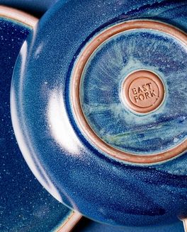 A shallow ceramic bowl upside down with a stamp that reads 'East Fork' and an unglazed circle. The bowl is a dappled deep glossy blue.