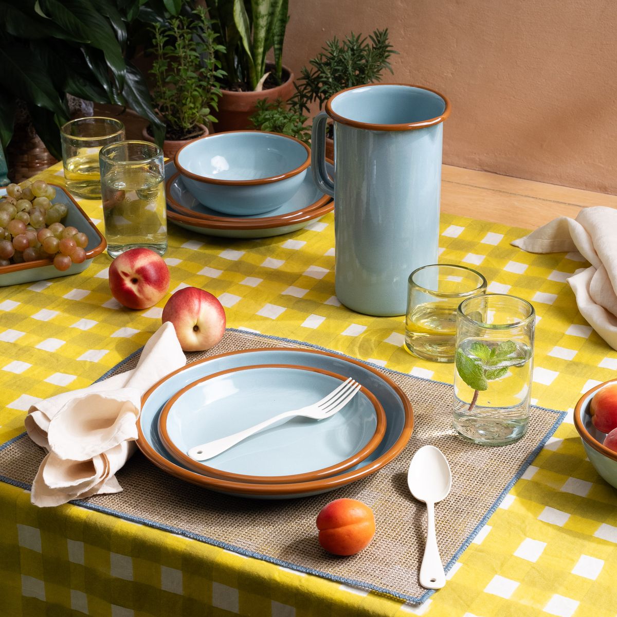 A table with a beautiful crosshatch yellow and white tablecloth, with light blue enamel plates, bowls, and pitcher, glassware and more.