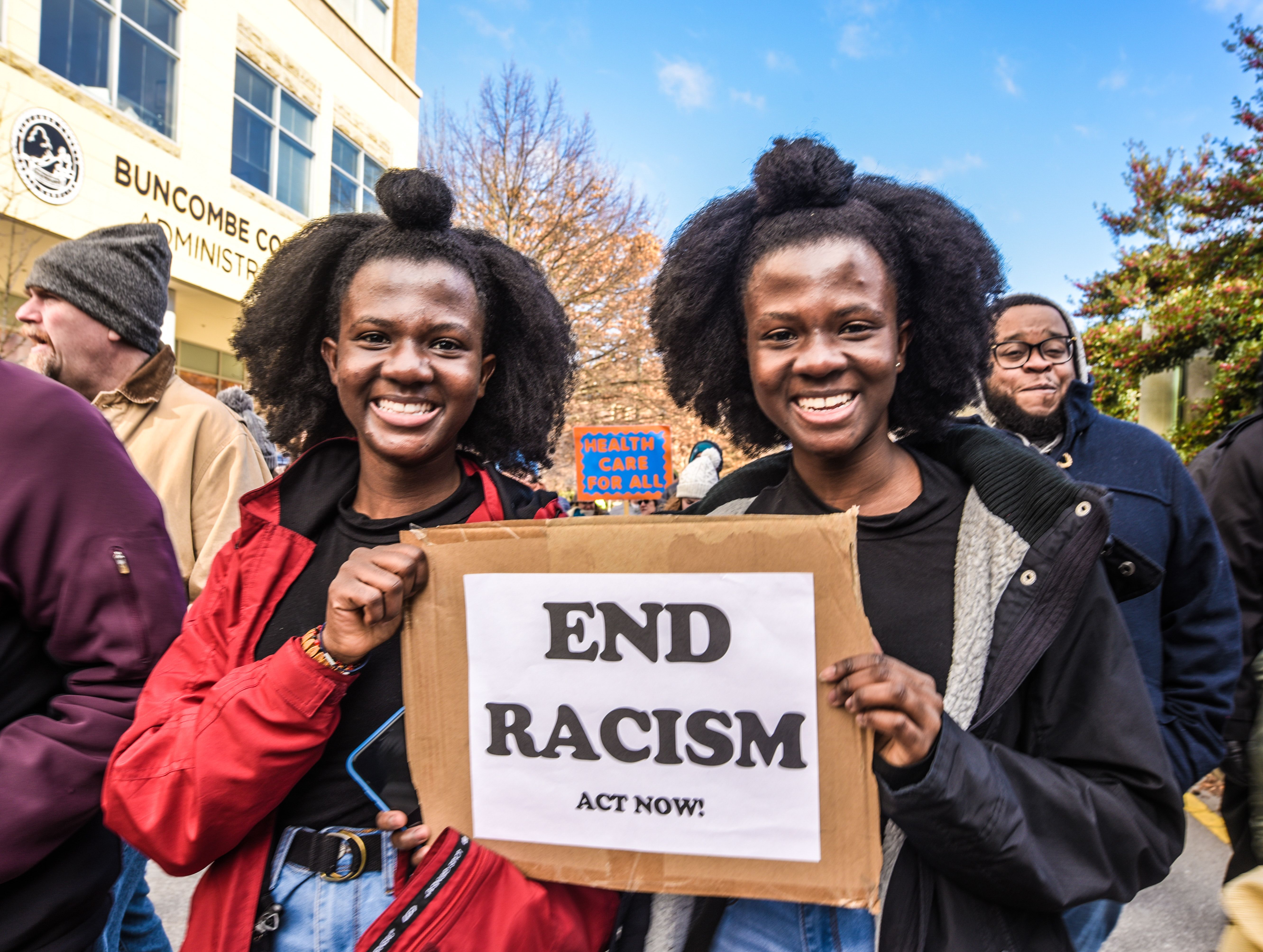 Twins are outside in a march and sharing a sign they both are holding that reads "End Racism - Act Now!"