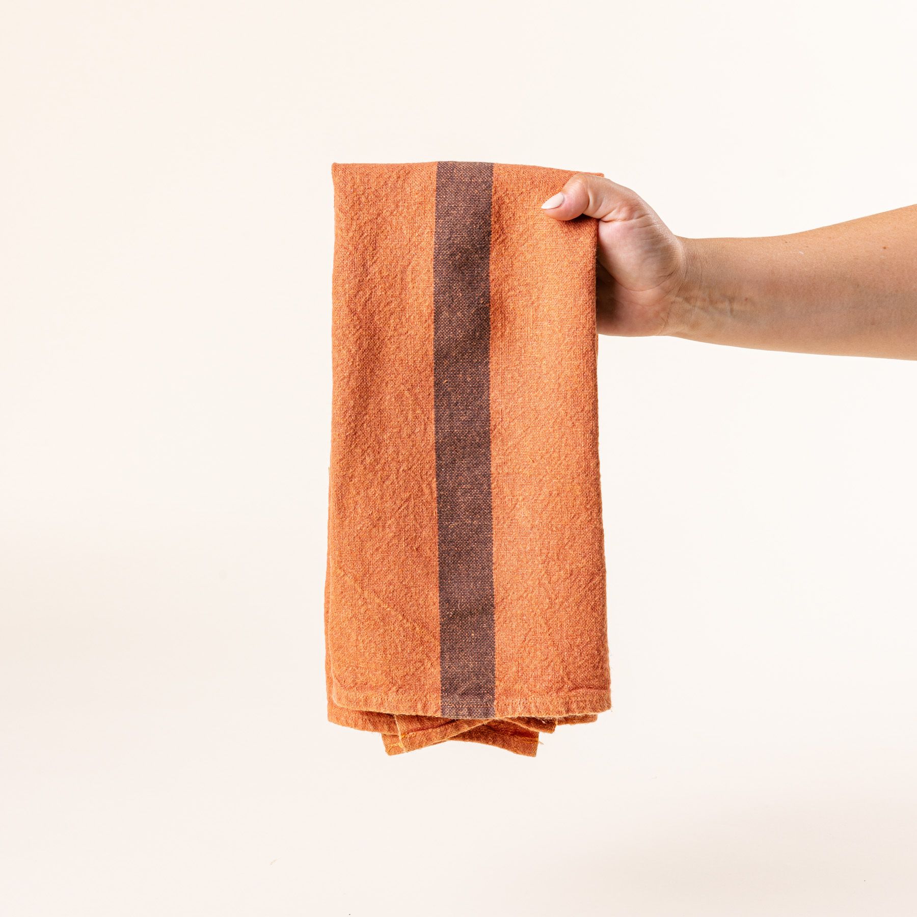 A hand holds a folded linen towel in a terracotta color with a plum stripe in the middle