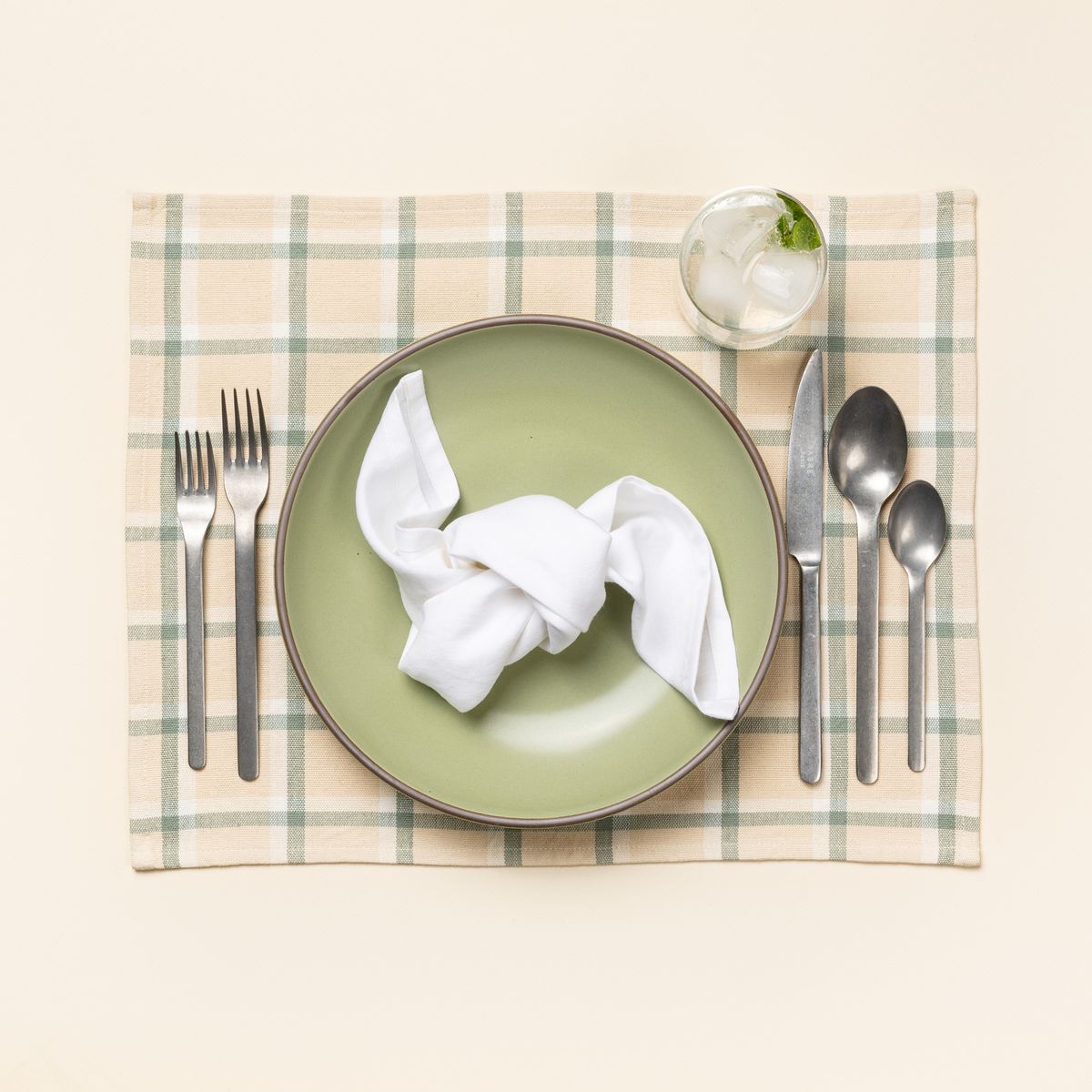 A table setting with a A gingham rectangular placemat in cream, off-white, and sage green colors, sage green plate, vintage steel flatware, and a folded white napkin