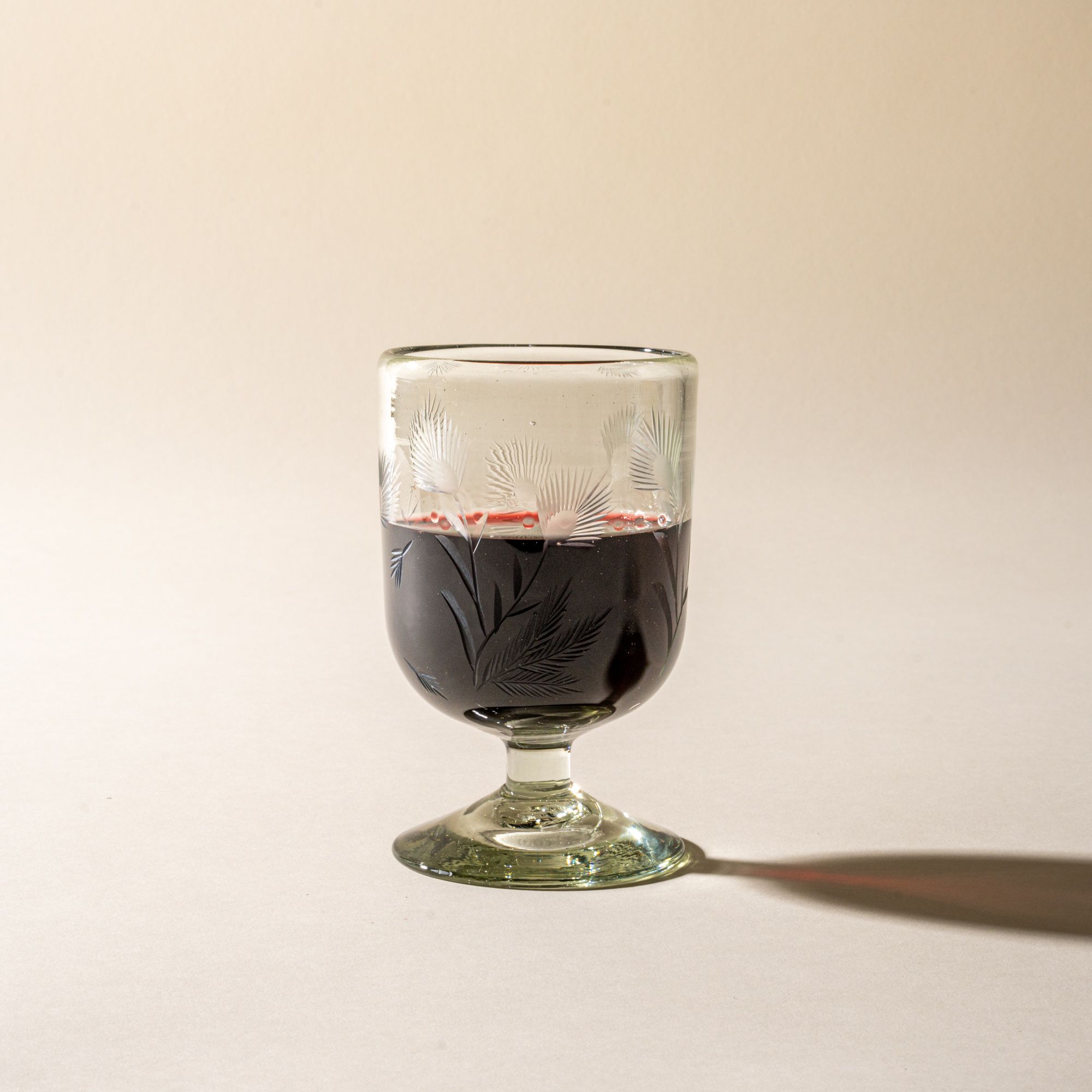 A clear short wine glass made of recycled glass with etched thistle designs on it, filled with red wine.